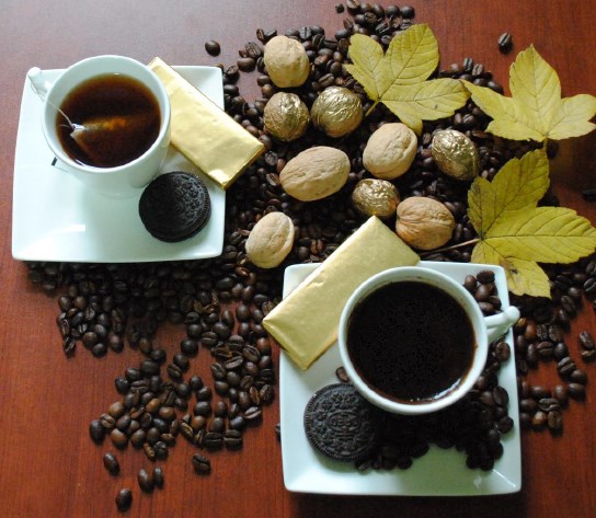 Zdjęcie przedstawiające 2 filiżanki z kawą i herbatą na podstawkach na których są ciastka a wokół rozsypane ziarna kawy, orzechy i żółte liście klonu