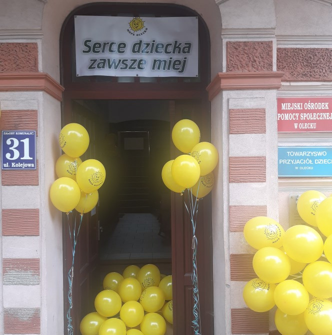 Wejście do Miejskiego Ośrodka Pomocy Społecznej w Olecku ozdobione żółtymi balonami a nad wejściem baner z napisem:"Serce dziecka zawsze miej"